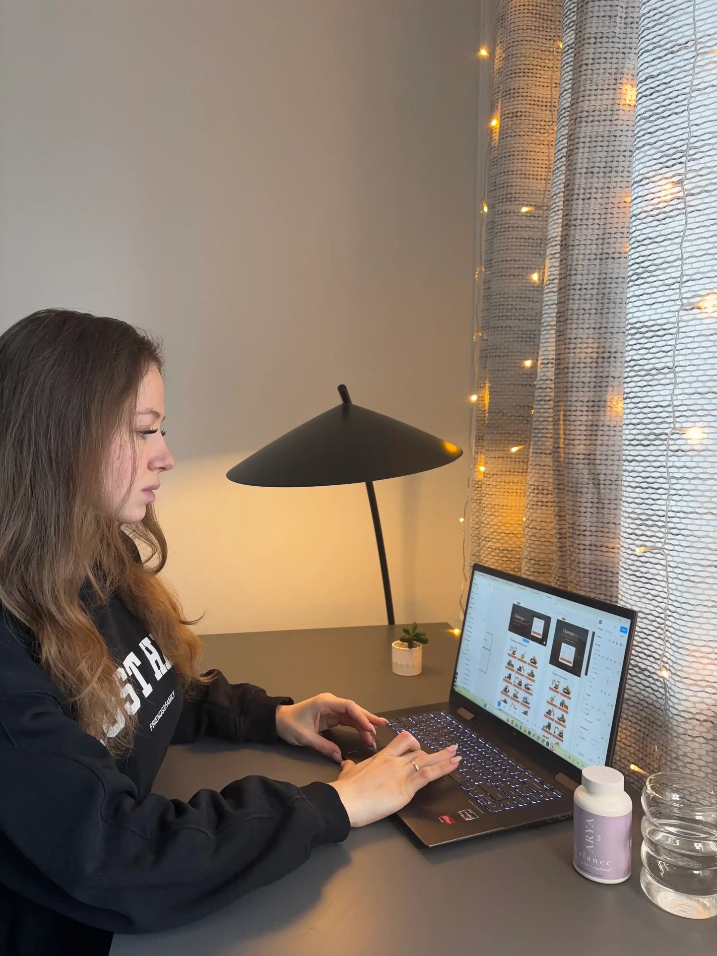 A beautiful lady using a laptop at a desk with a bottle of Balance by ARYA Lab, a lamp and decorative lights in the background