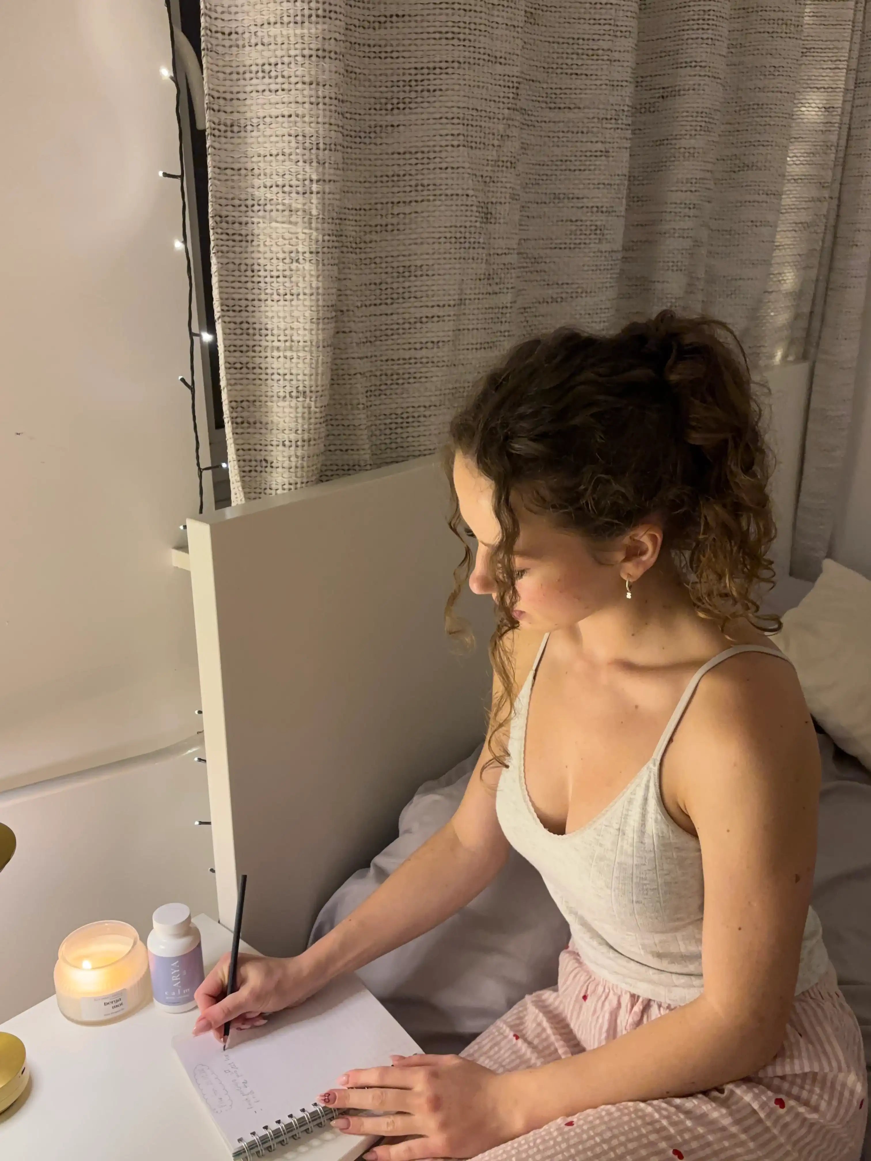 Woman sitting on a bed writing in a notebook with a lit candle and bottle of Calm by ARYA Lab nearby.