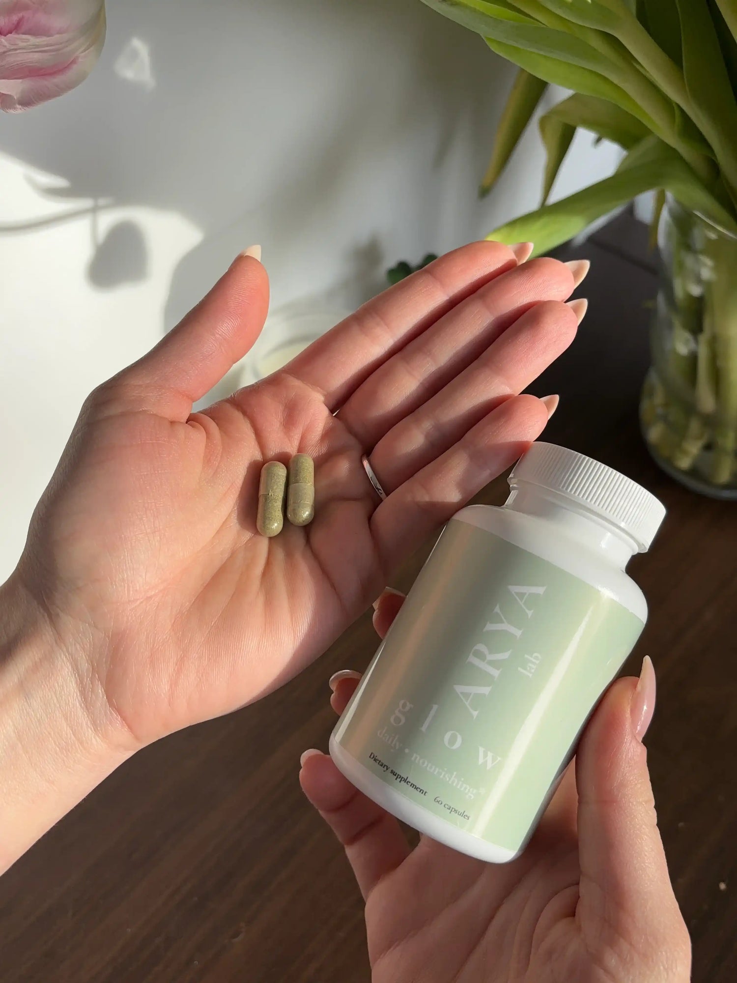 Hand holding two capsules and a supplement bottle (Glow by ARYA Lab) labeled 'Arya' on a wooden surface with flowers in the background.