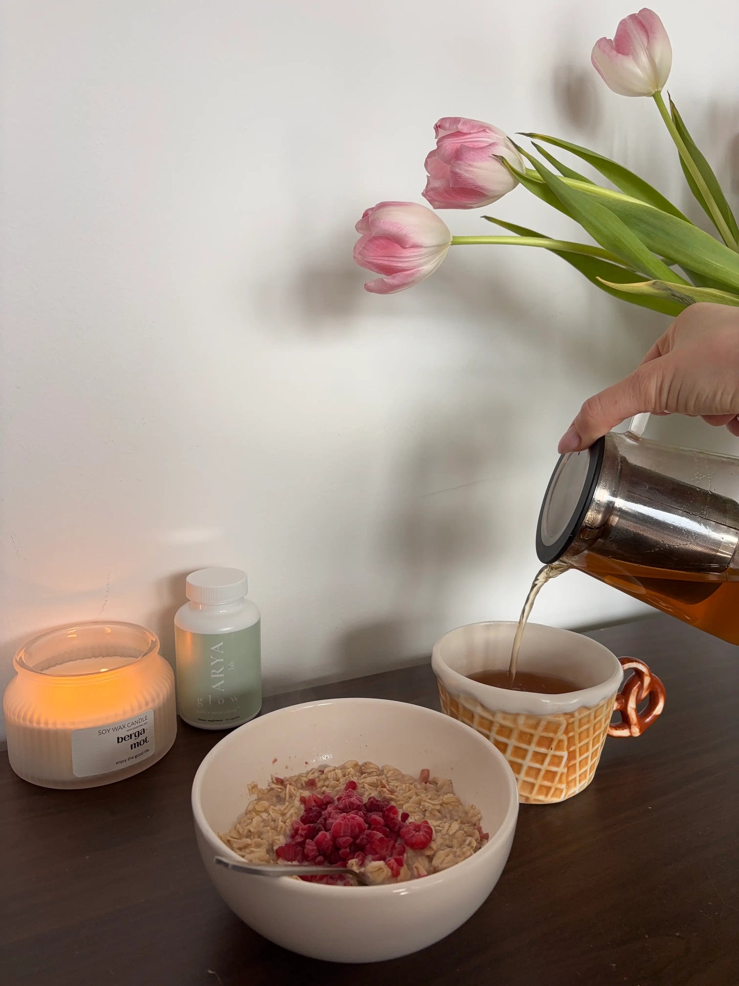 Person pouring tea into a cup with a bowl of cereal and fruit, candle, a bottle of Glow and flowers on a table.